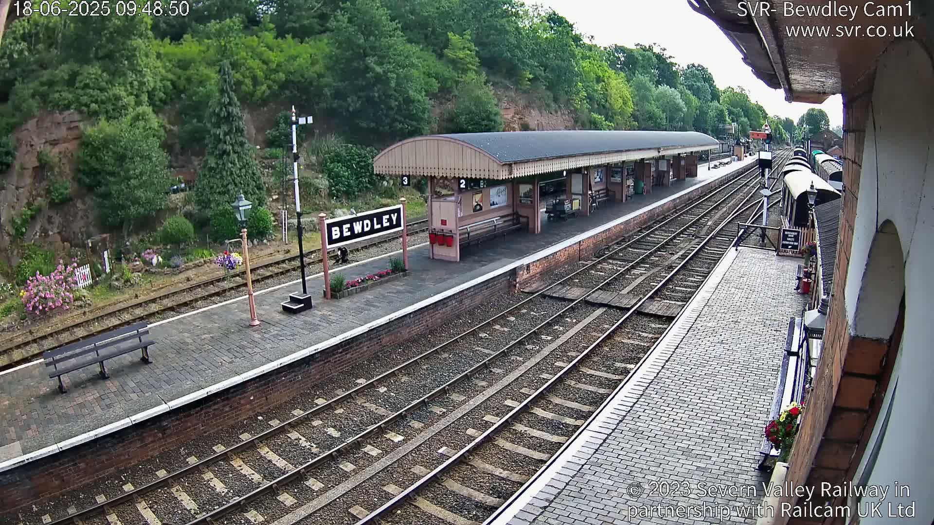 A train station platform with a covered waiting area, multiple train tracks, and lush green hillsides in the background on a partly cloudy day.