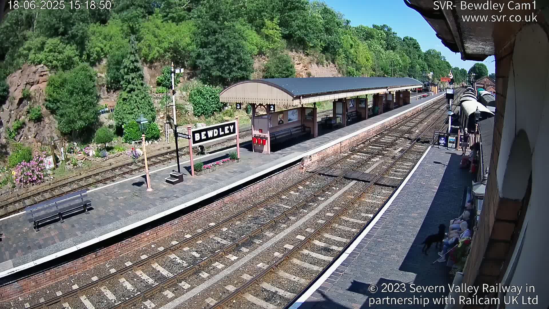 A train station platform on a sunny day with multiple train tracks, a covered waiting area, and several people sitting on benches.