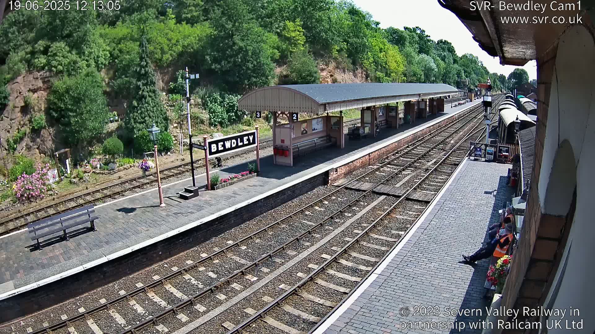 A sunny view of a train station platform with multiple train tracks, a covered waiting area, and a few people sitting near the edge of the platform.