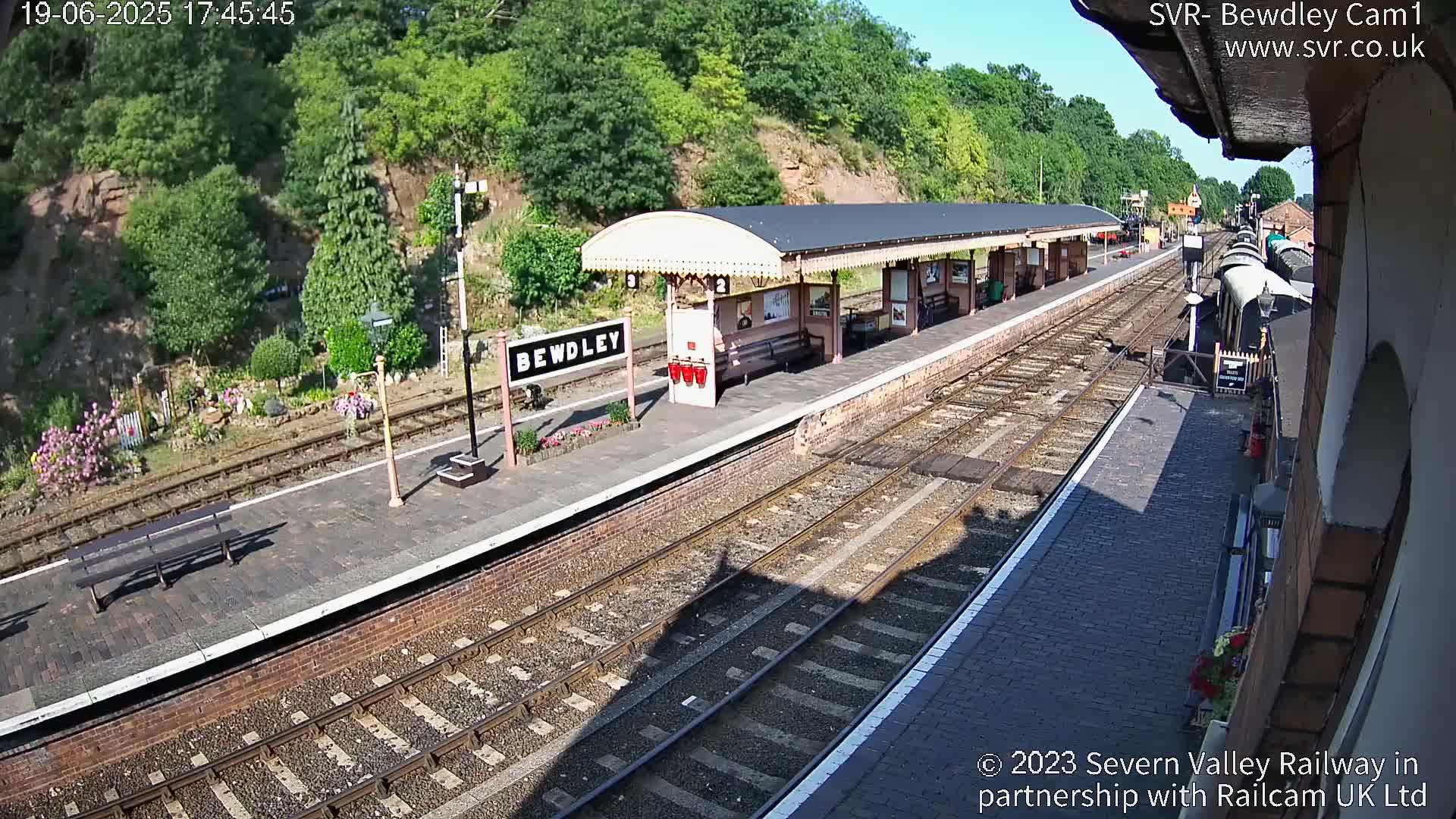 A sunny view of a train station platform with several train tracks and a covered waiting area, backed by a hillside covered in lush green vegetation.
