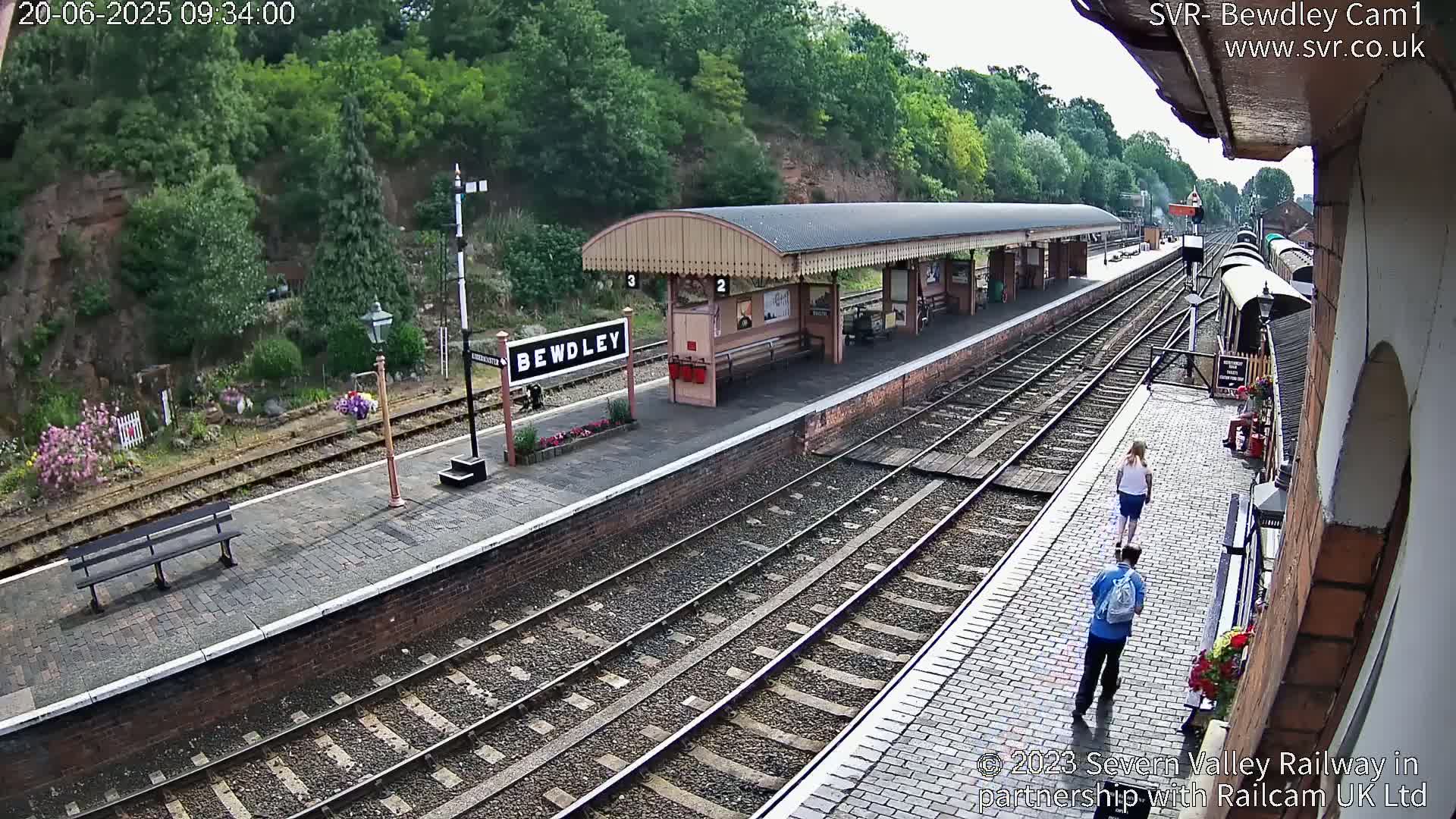 A high-angle view of a train station platform with two people walking on the platform under a partly sunny sky, with railway tracks and greenery visible in the background.