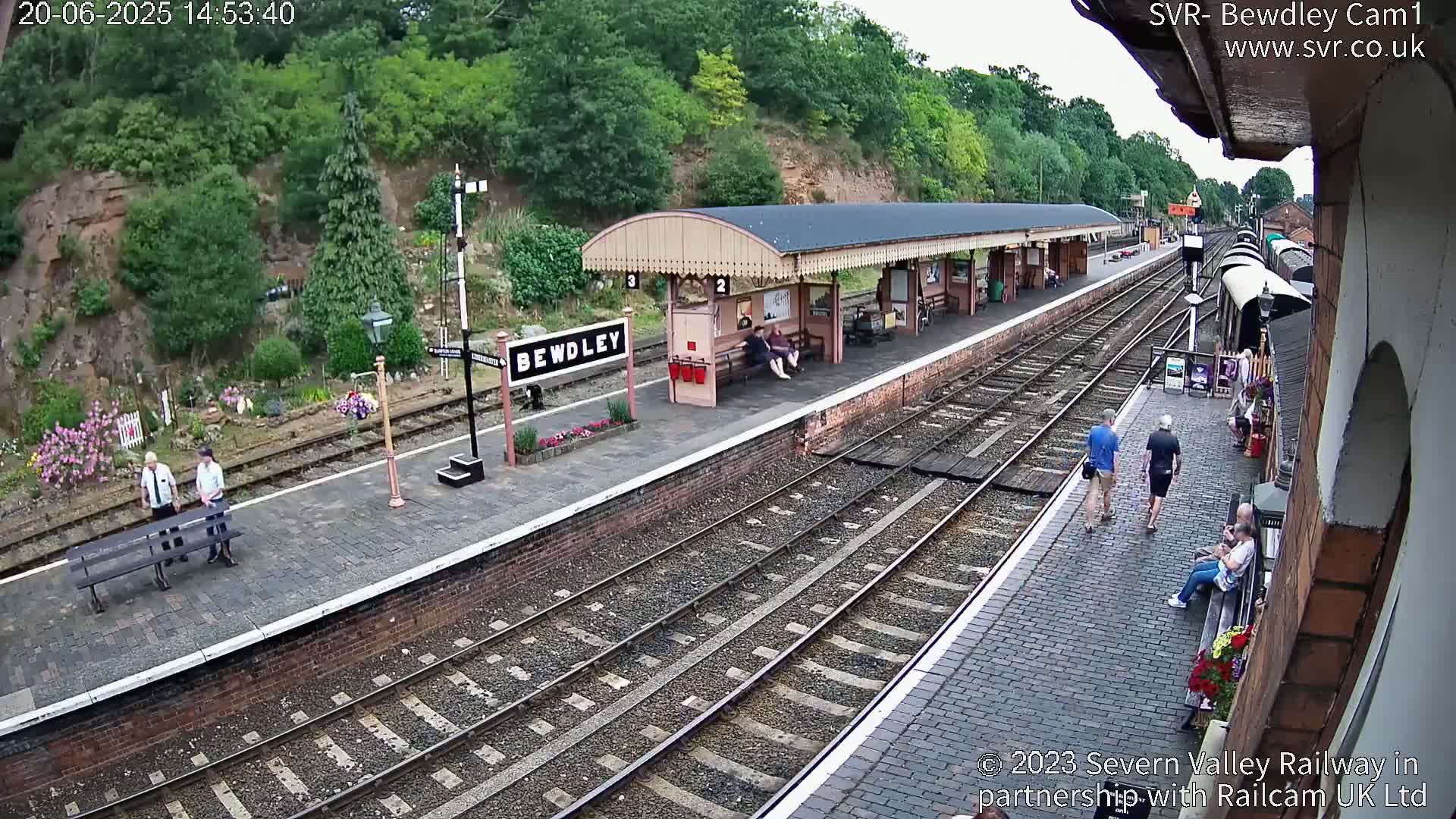 A train station platform with several people, some seated and some walking, on a sunny day, next to train tracks and a wooded hillside.