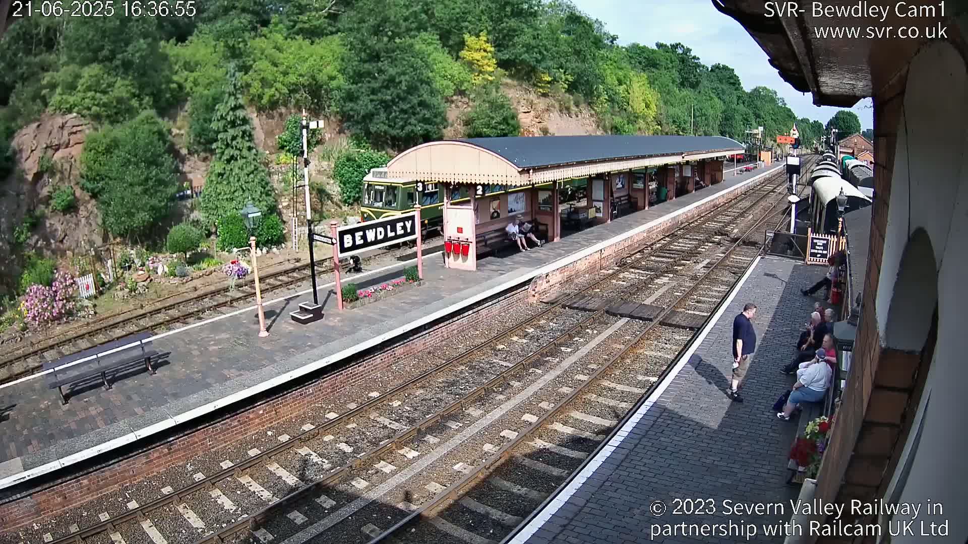 A train station platform on a sunny day, with a train at the platform, several people sitting and standing on the platform, and greenery visible beyond the tracks.