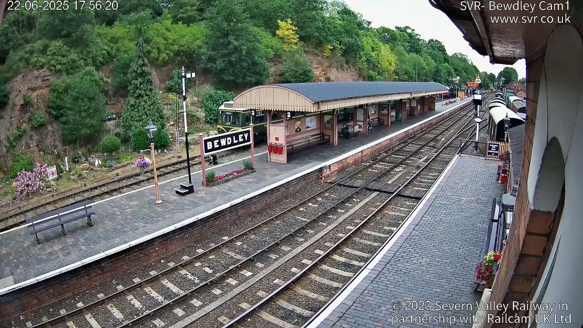 A train sits at a station platform next to a covered waiting area, under a partly sunny sky.
