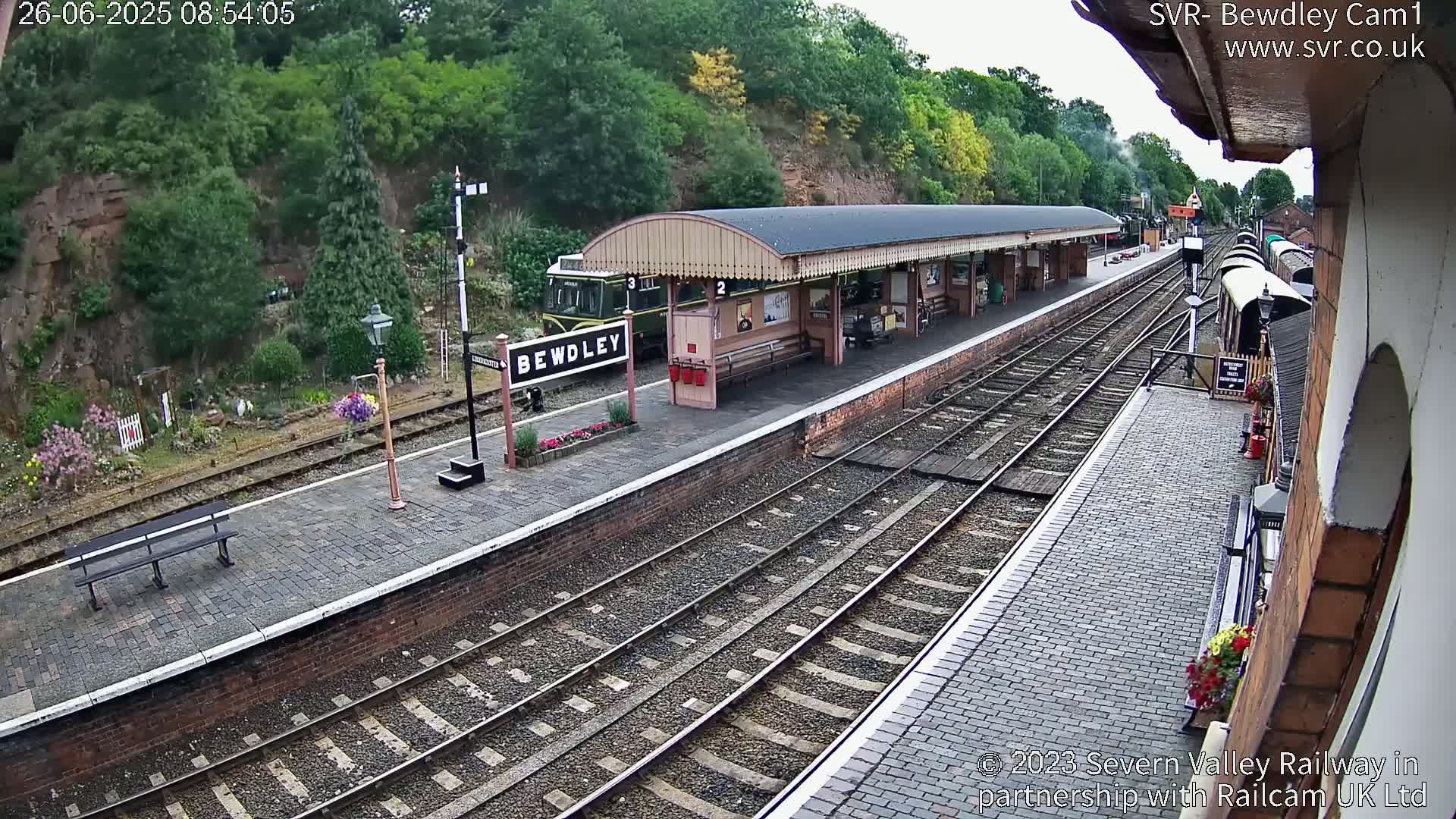 A train sits at a railway station platform next to a covered waiting area under a partly cloudy sky.