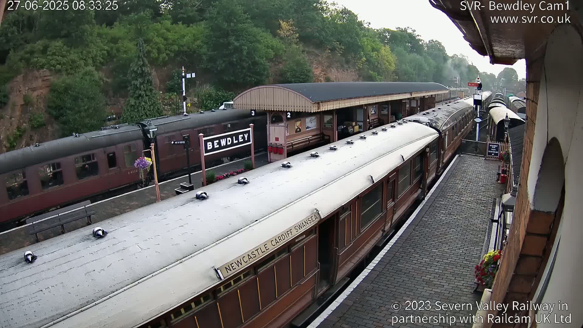 A long, brown passenger train sits at a station next to another train partially obscured by a covered platform, under an overcast sky.