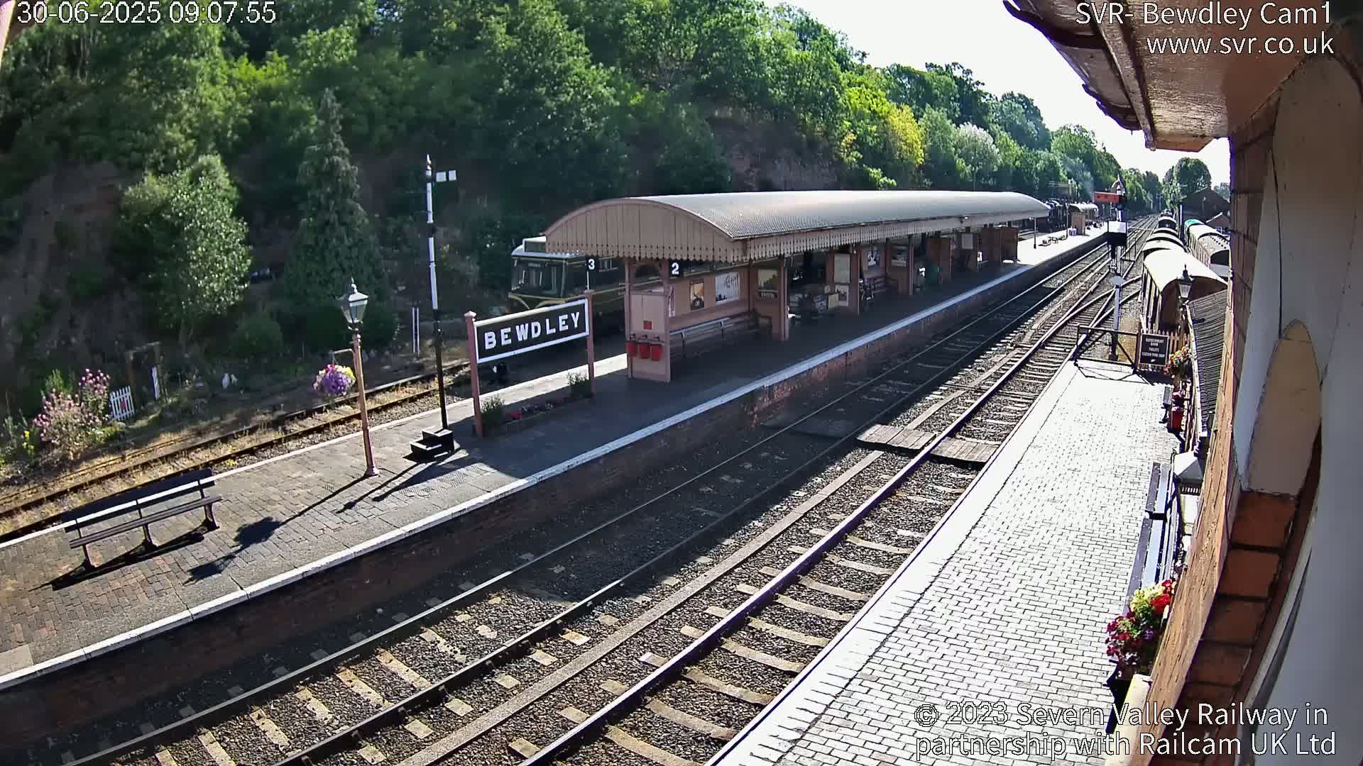 A sunny day at a train station shows a platform with a covered waiting area, railway tracks extending into the distance, and a train partially visible.