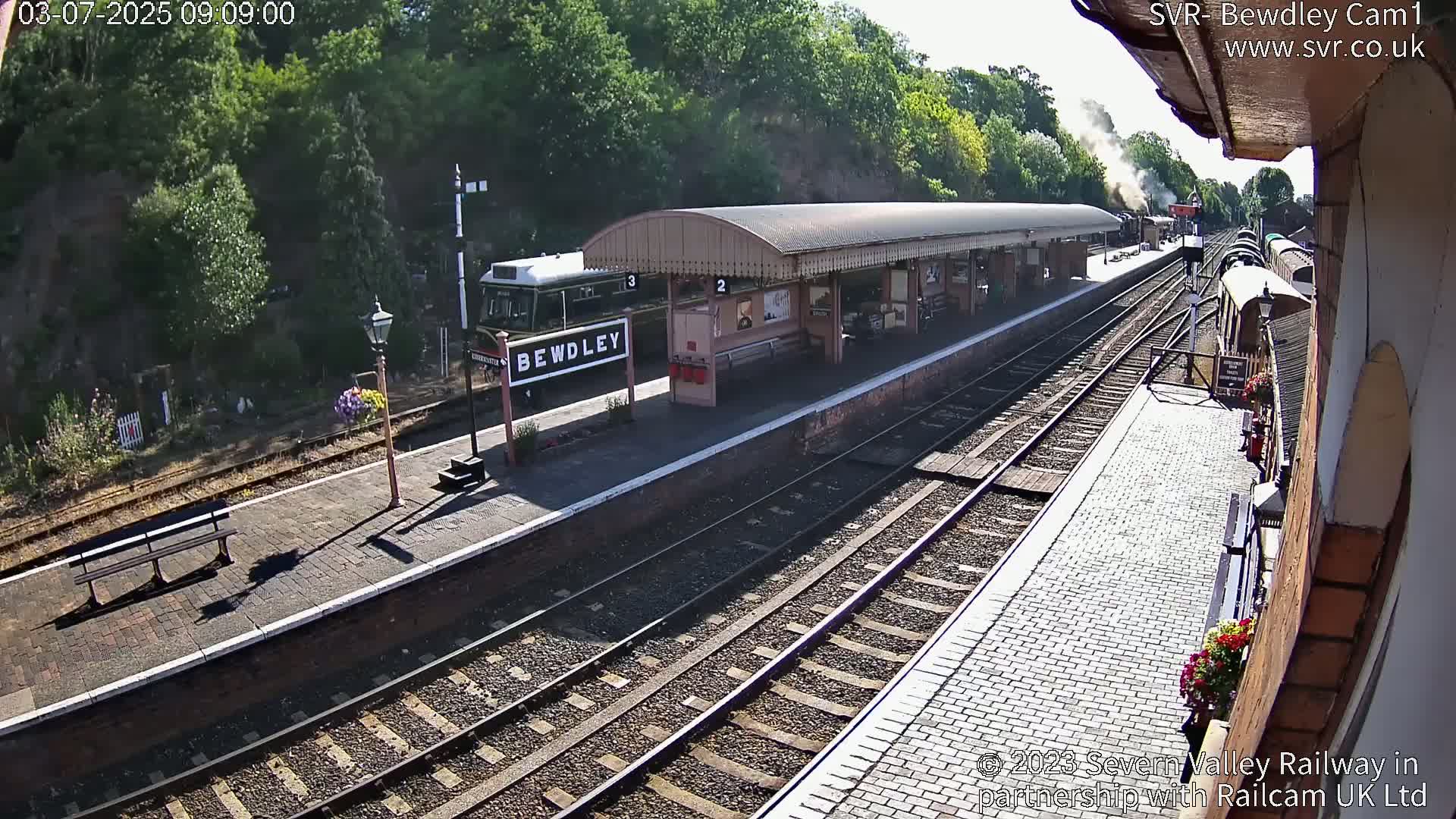 A sunny view of a train station platform with a train stopped at the platform,  multiple train tracks, and a covered waiting area.