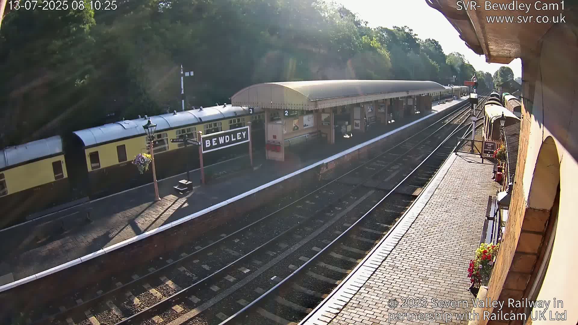 A sunny day at a train station shows passenger train cars at the platform, multiple train tracks, and a brick building.