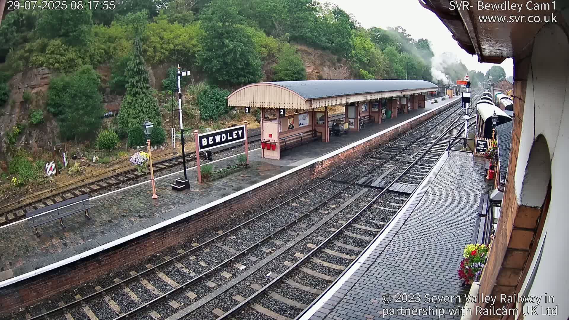 A wet train station platform with a covered waiting area, train tracks, and a steam train partially visible in the distance, surrounded by greenery on a hillside.