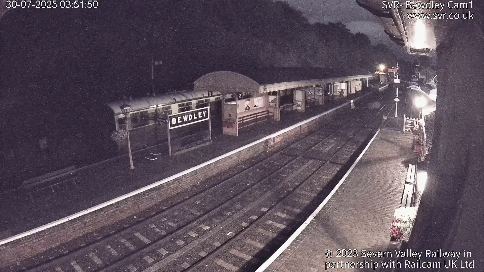 A wet train station platform with a covered waiting area, train tracks, and a steam train partially visible in the distance, surrounded by greenery on a hillside.
