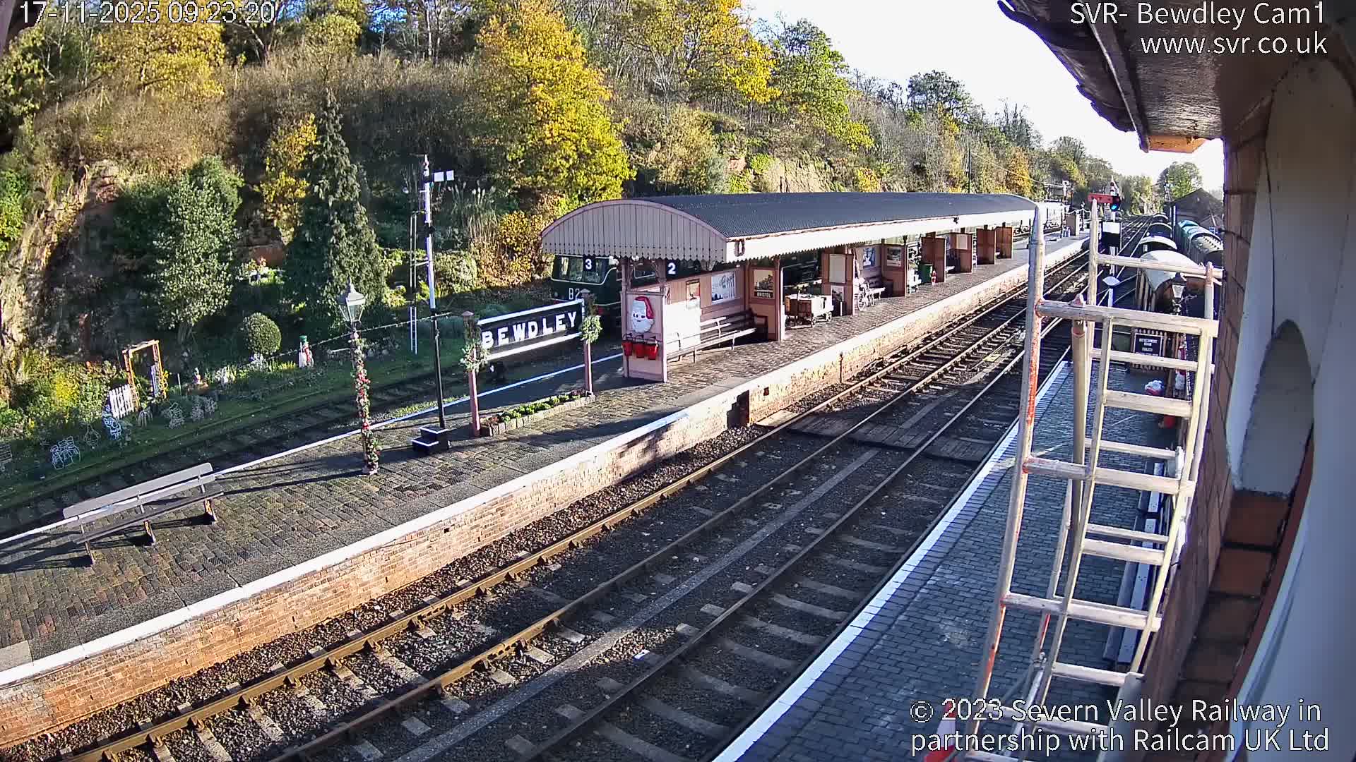 A sunny, elevated view reveals the charming Bewdley train station, adorned with festive decorations, featuring a long covered platform and multiple tracks, with scaffolding visible on the right and a backdrop of vibrant autumn trees.