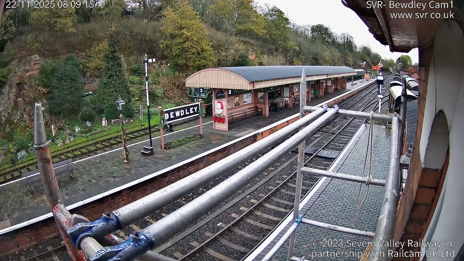 An outdoor view of Bewdley train station reveals a long platform building with a green train carriage, tracks, and festive decorations, partially obscured by scaffolding in the foreground, all under an overcast sky.