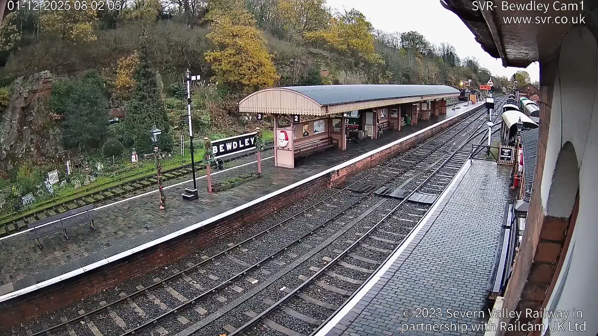 A historic railway station adorned with festive Christmas decorations stands under an overcast sky on a wet day, featuring multiple tracks, brick platforms, a covered waiting area, and a wooded hillside in the background.