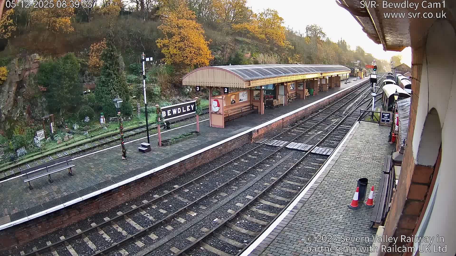 A sunny, clear morning reveals a festive Bewdley train station, featuring brick platforms, multiple tracks, and Christmas decorations amidst autumnal trees on a hillside.