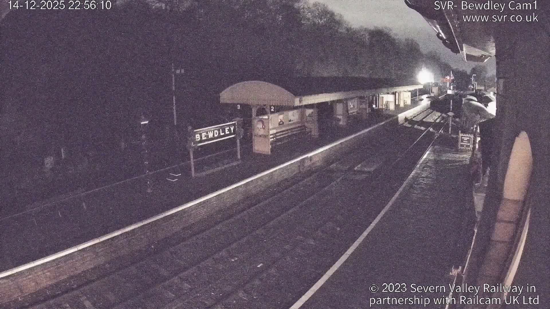 A sunny, clear morning reveals a festive Bewdley train station, featuring brick platforms, multiple tracks, and Christmas decorations amidst autumnal trees on a hillside.