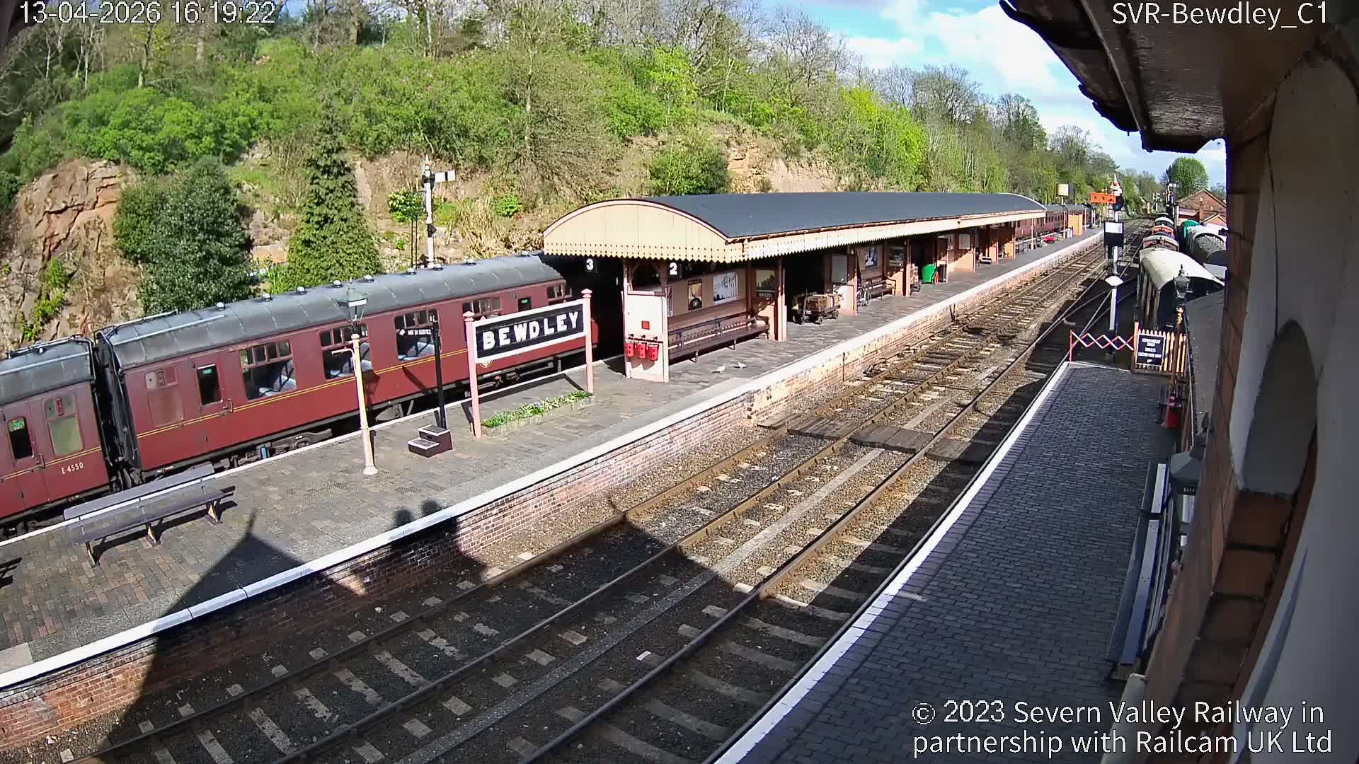 A sunny, clear morning reveals a festive Bewdley train station, featuring brick platforms, multiple tracks, and Christmas decorations amidst autumnal trees on a hillside.