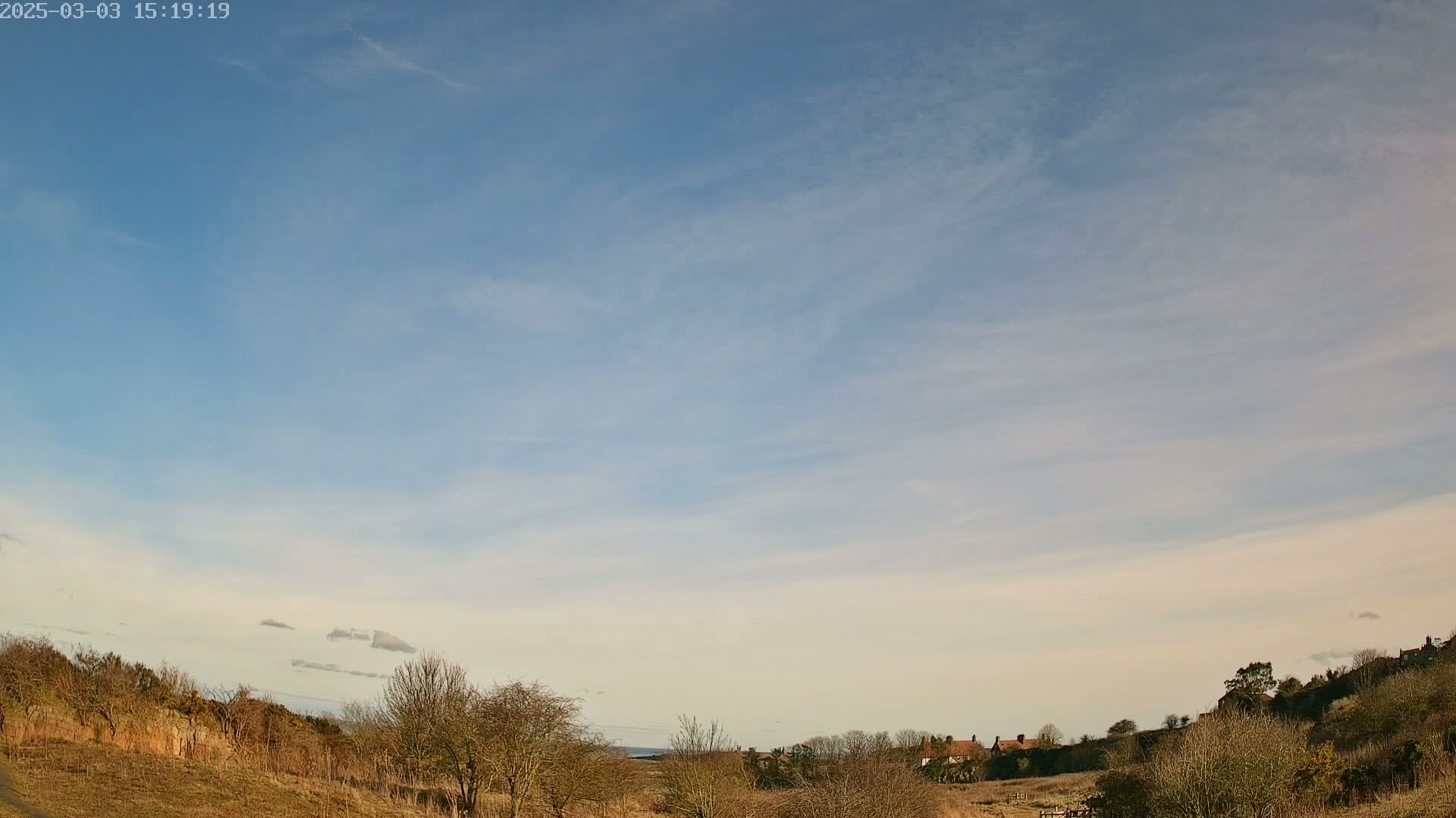 A mostly clear, light blue sky stretches over a sparsely vegetated landscape with a few buildings visible in the distance.