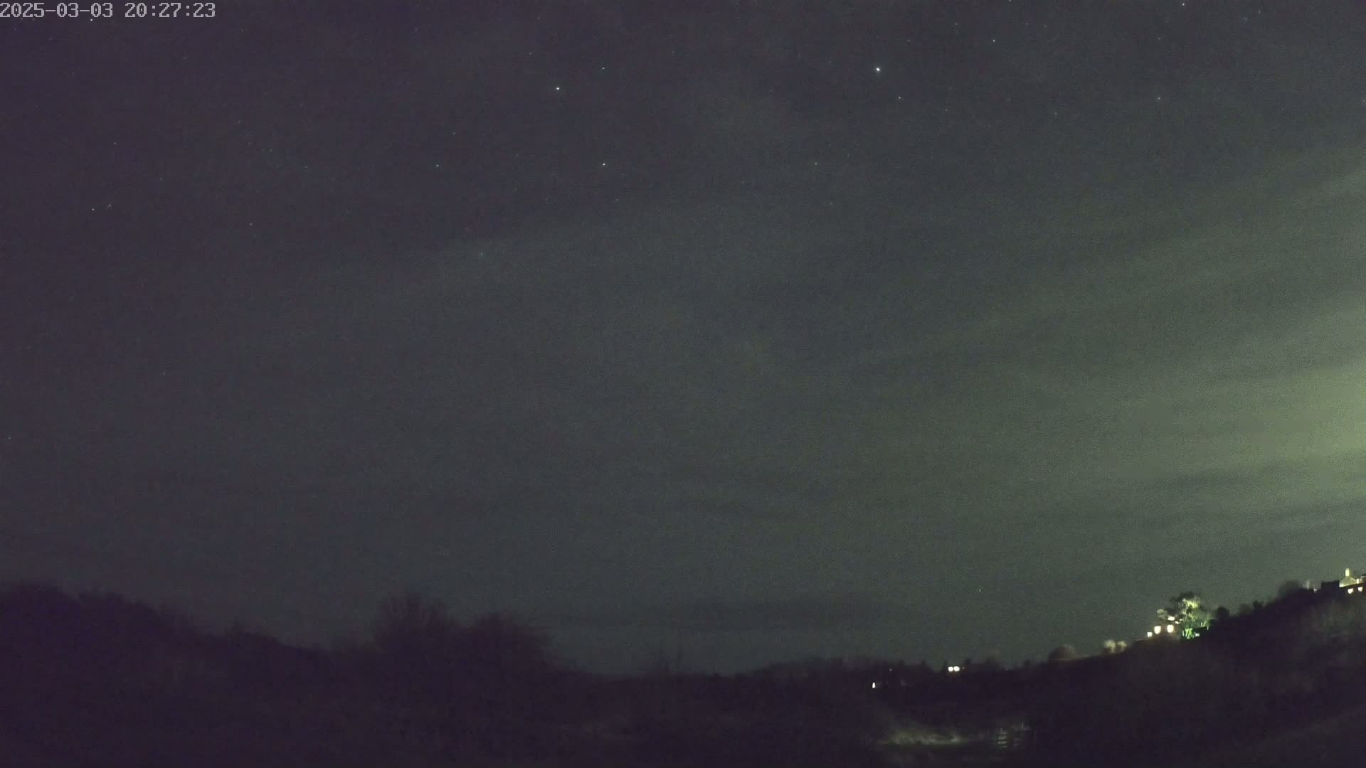 A night sky with some stars and thin clouds above a dark, silhouetted landscape.