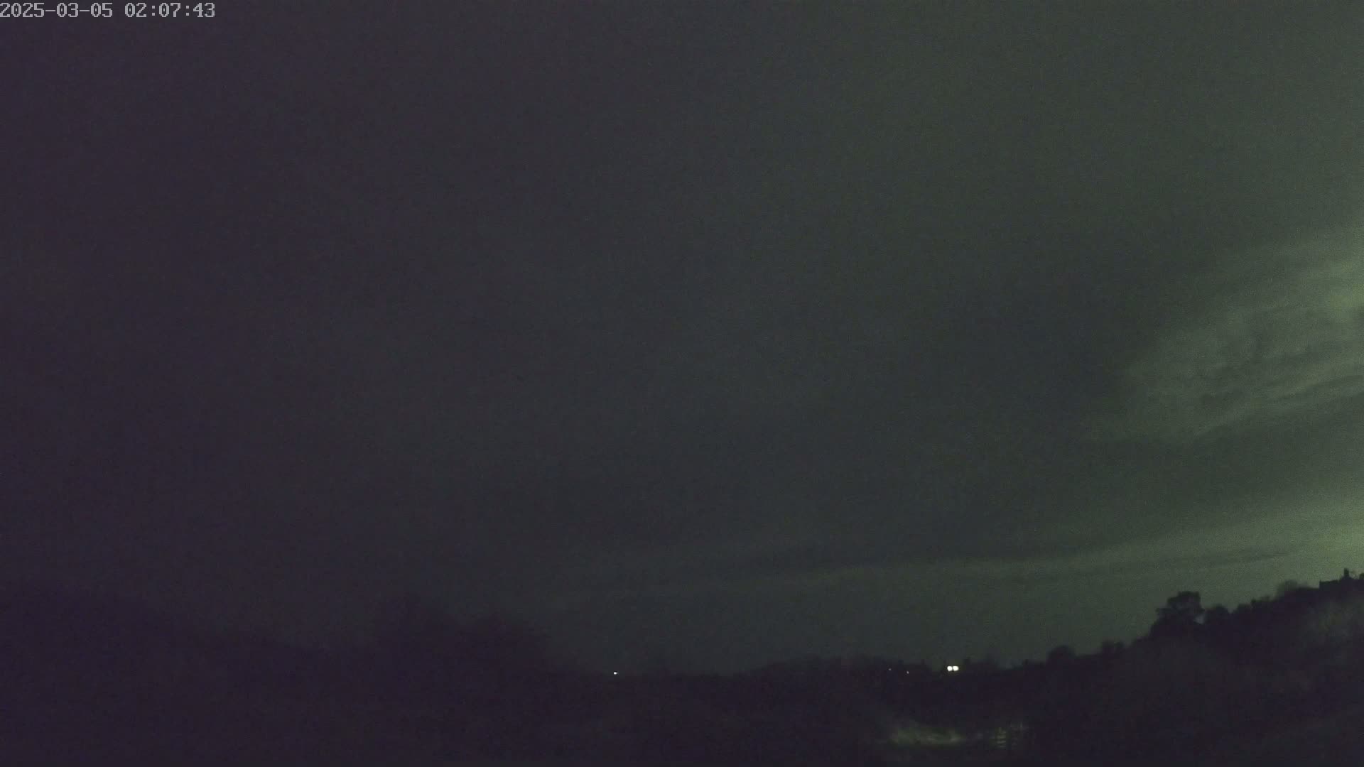 A dark, overcast night sky hangs over a silhouetted landscape of trees and low hills.