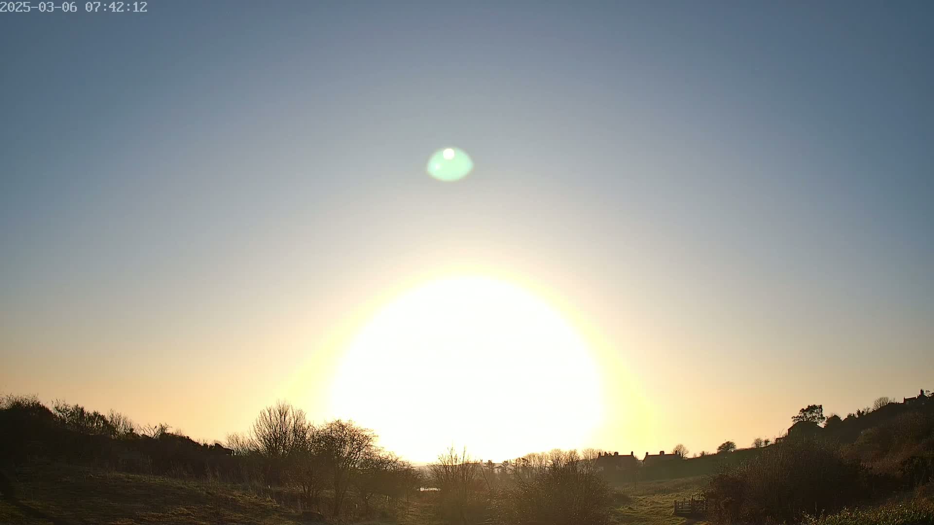 A bright sun shines over a mostly clear sky behind a silhouetted landscape of trees and buildings.