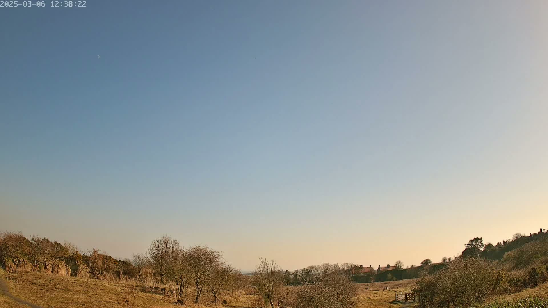 A mostly clear, pale blue sky with a sliver of a crescent moon above a hilly landscape of dry, brown grass and leafless bushes, with a few distant houses visible on the horizon.