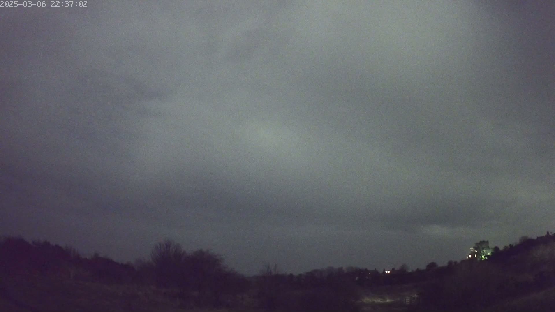 A dark, overcast night sky looms over a silhouetted landscape of trees and low hills.