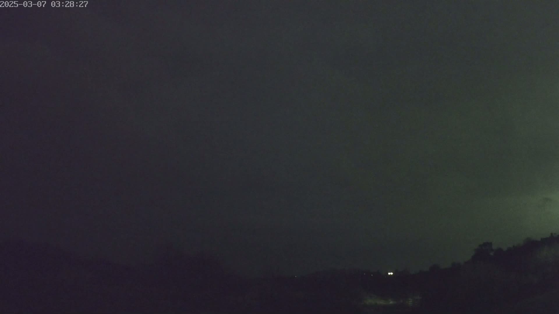 A dark, overcast night sky looms over a silhouetted landscape of trees and distant lights.