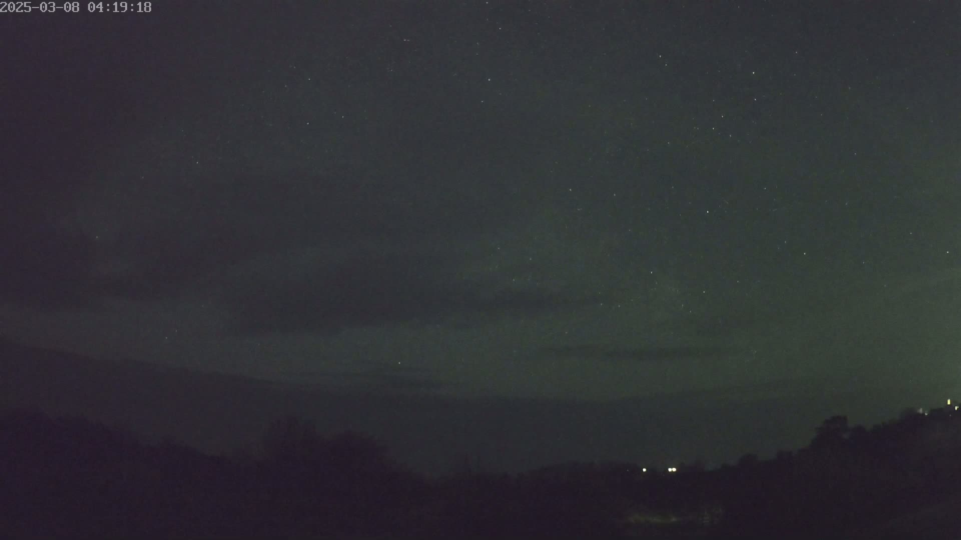 A nighttime landscape shows a starry sky with some clouds above a dark, silhouetted line of trees and hills.