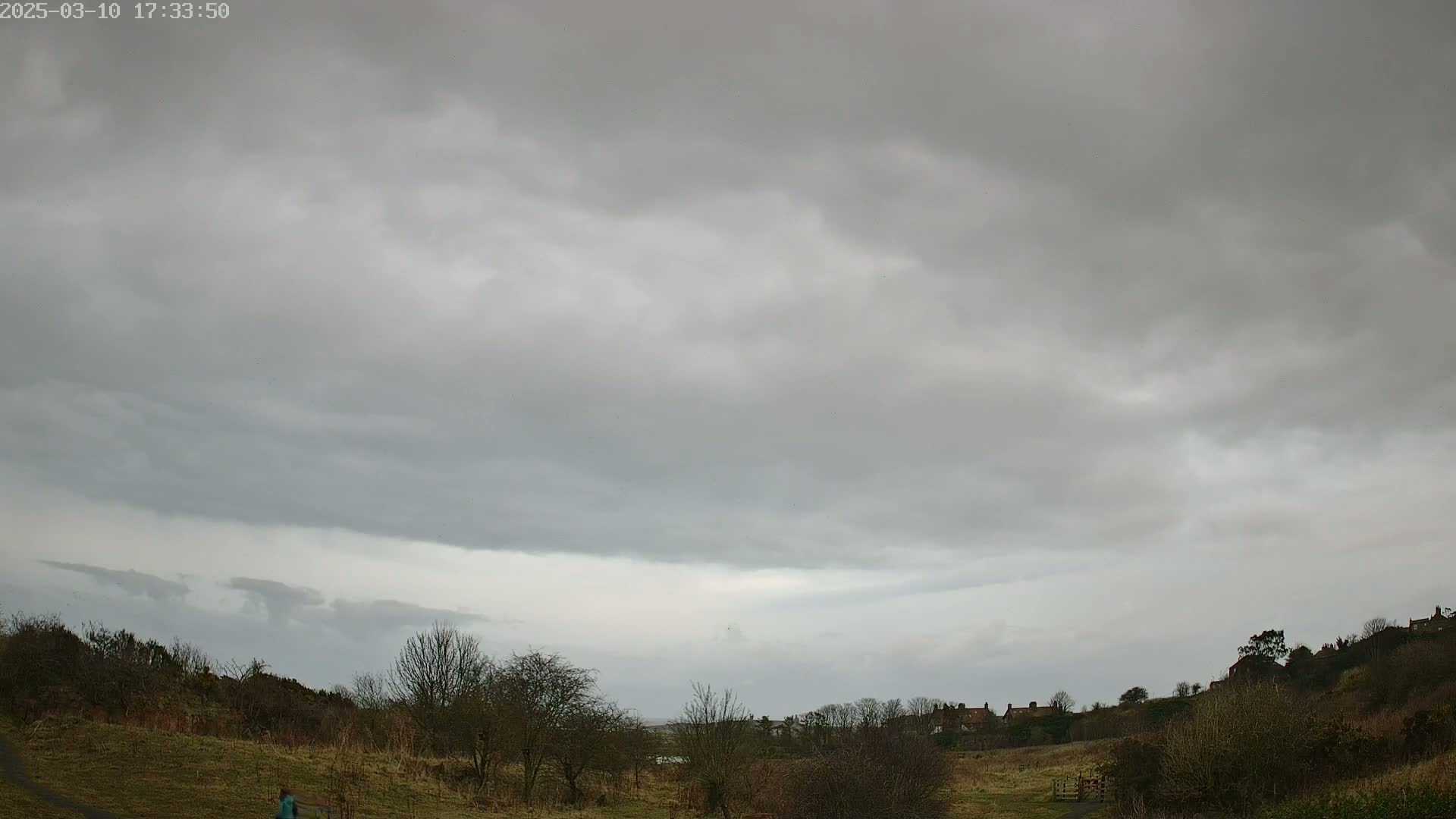 A person walks along a path through a mostly barren, hilly landscape under a cloudy, overcast sky.