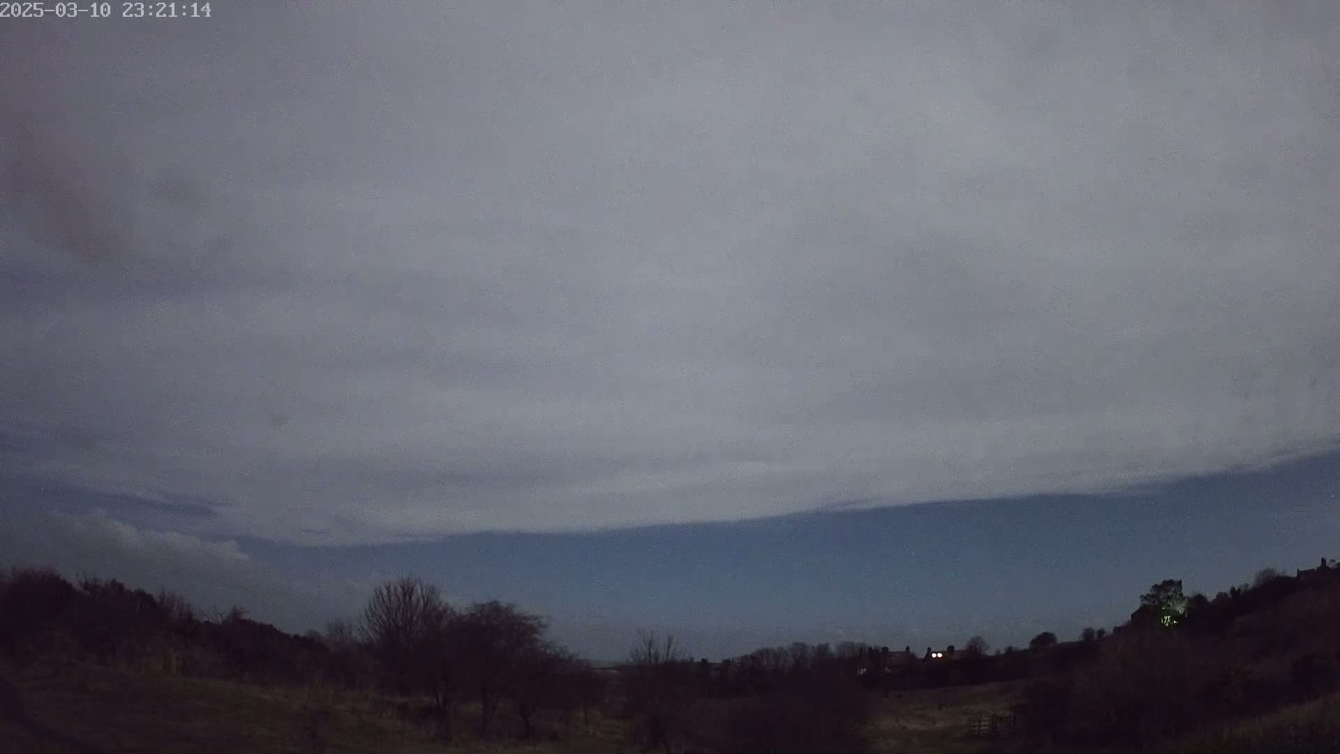 Under a mostly cloudy, dark night sky, a landscape of dark, silhouetted trees and a few distant lights is visible.