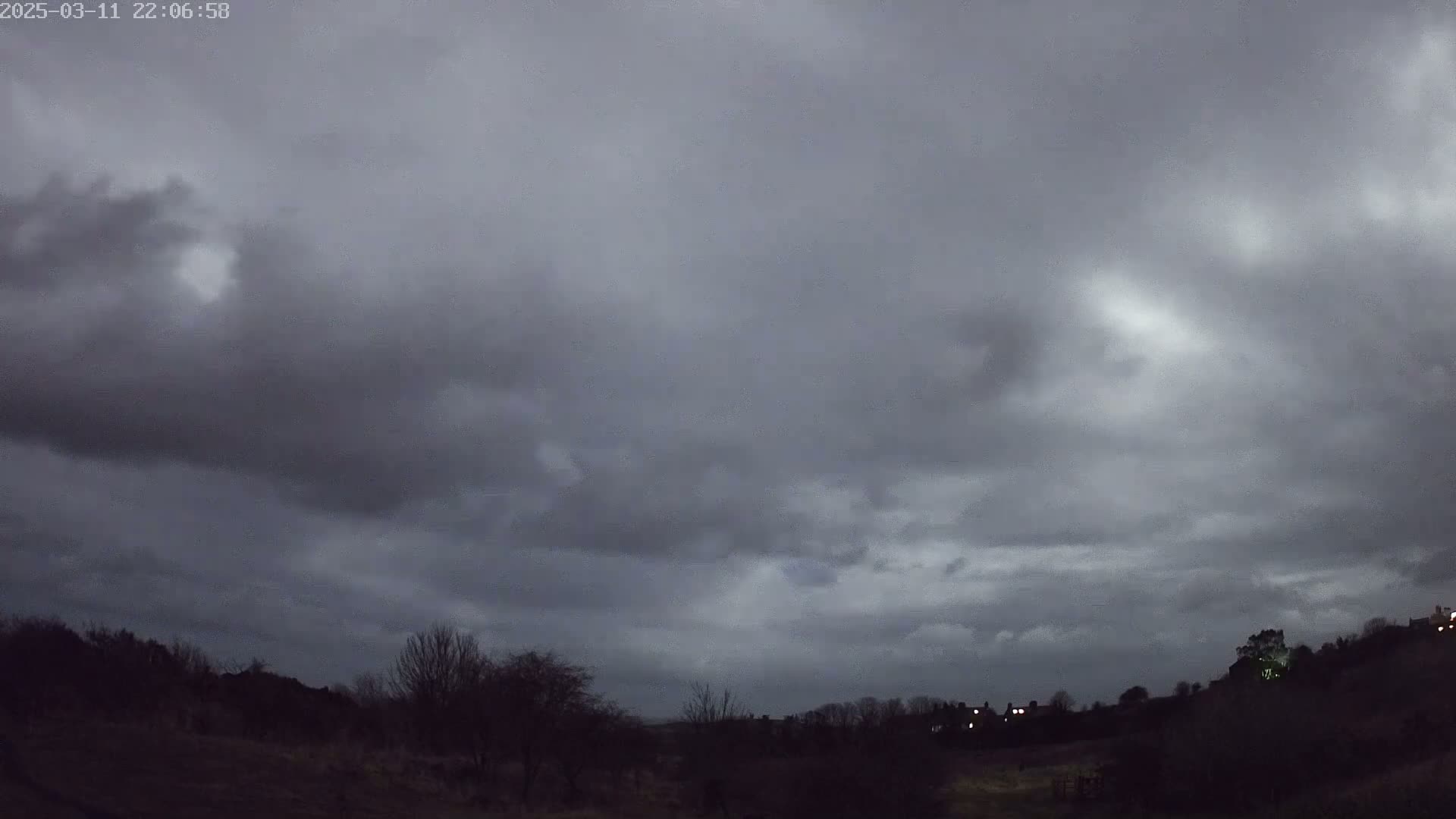 A dark, overcast sky hangs over a silhouetted landscape of trees and buildings at night.