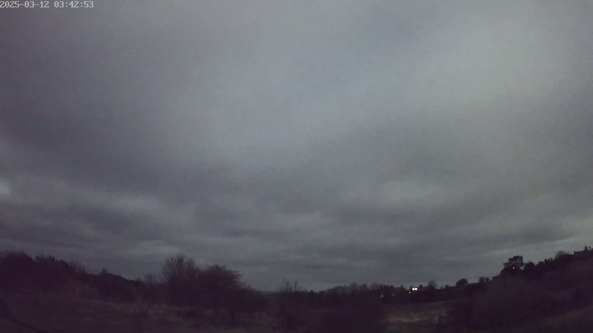 A dark, overcast sky hangs over a silhouetted landscape of low hills and trees at night.