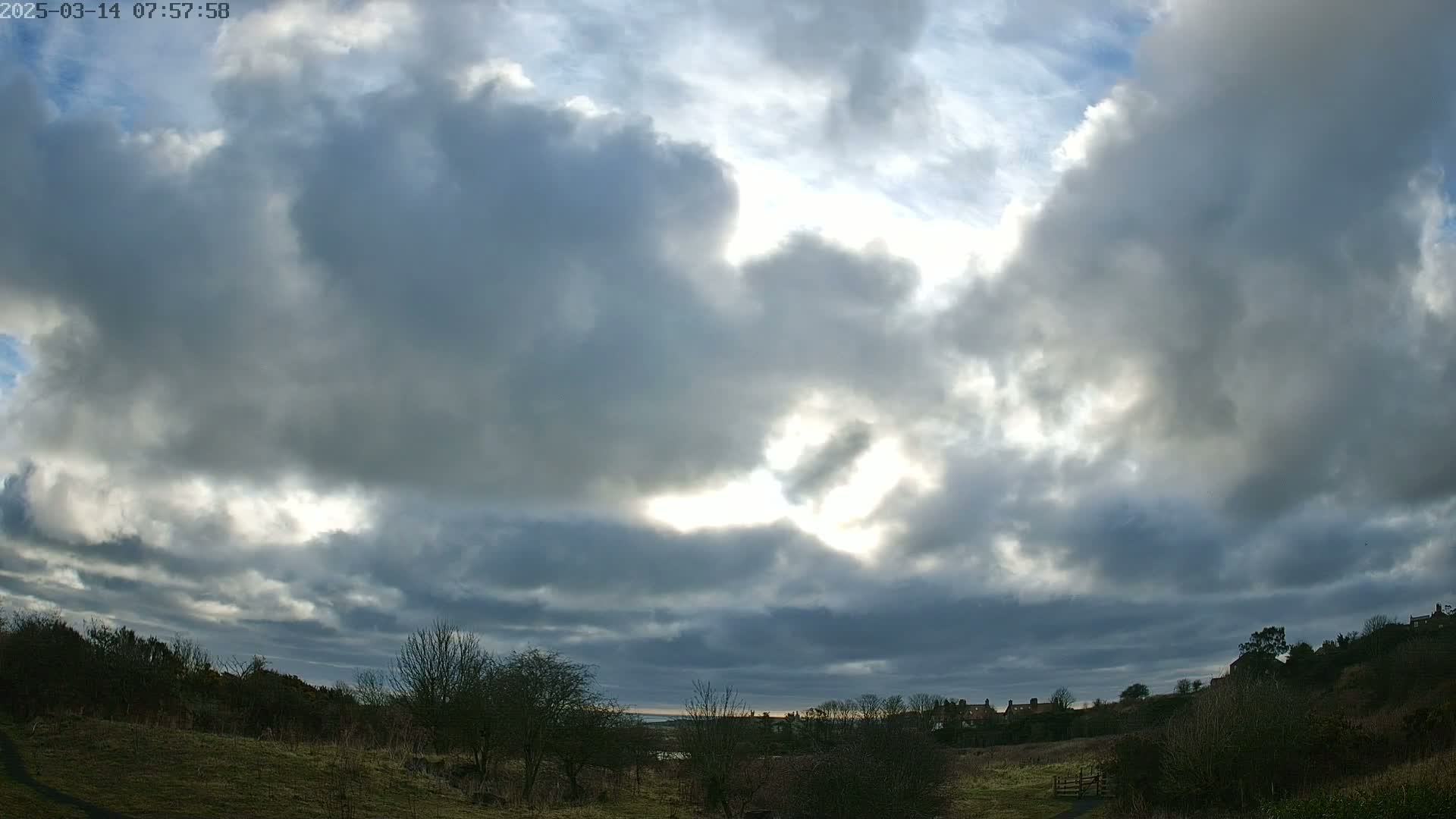 A mostly cloudy sky with breaks of sunlight illuminates a grassy field and distant houses under a mostly overcast sky.