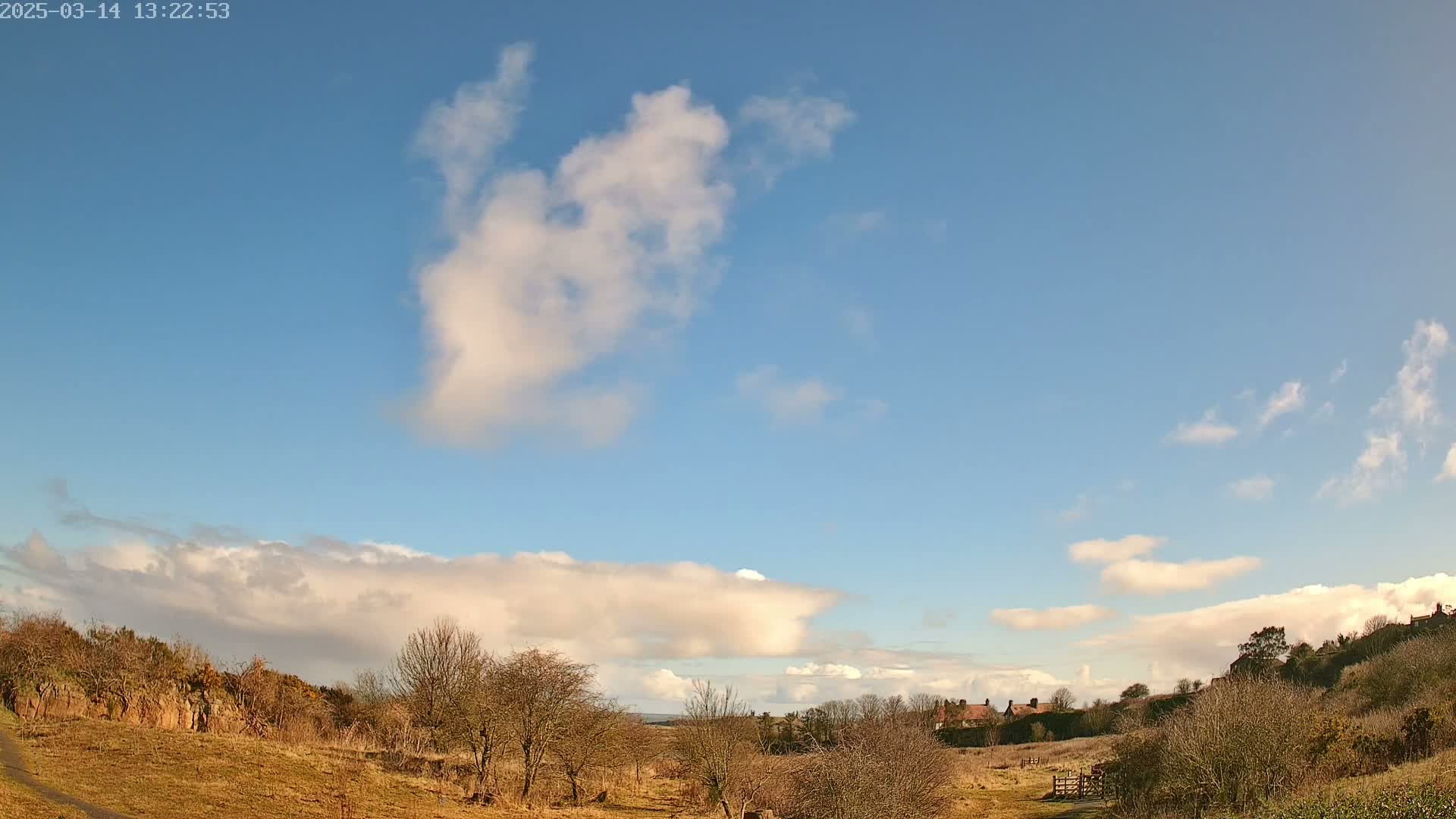 A mostly sunny day with scattered clouds over a brown, sparsely vegetated landscape with a few buildings in the distance.