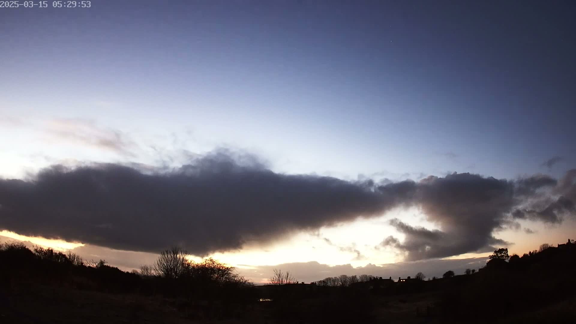 A low-lying, dark cloud formation obscures a sunset over a silhouetted landscape of trees and buildings under a mostly clear, twilight sky.