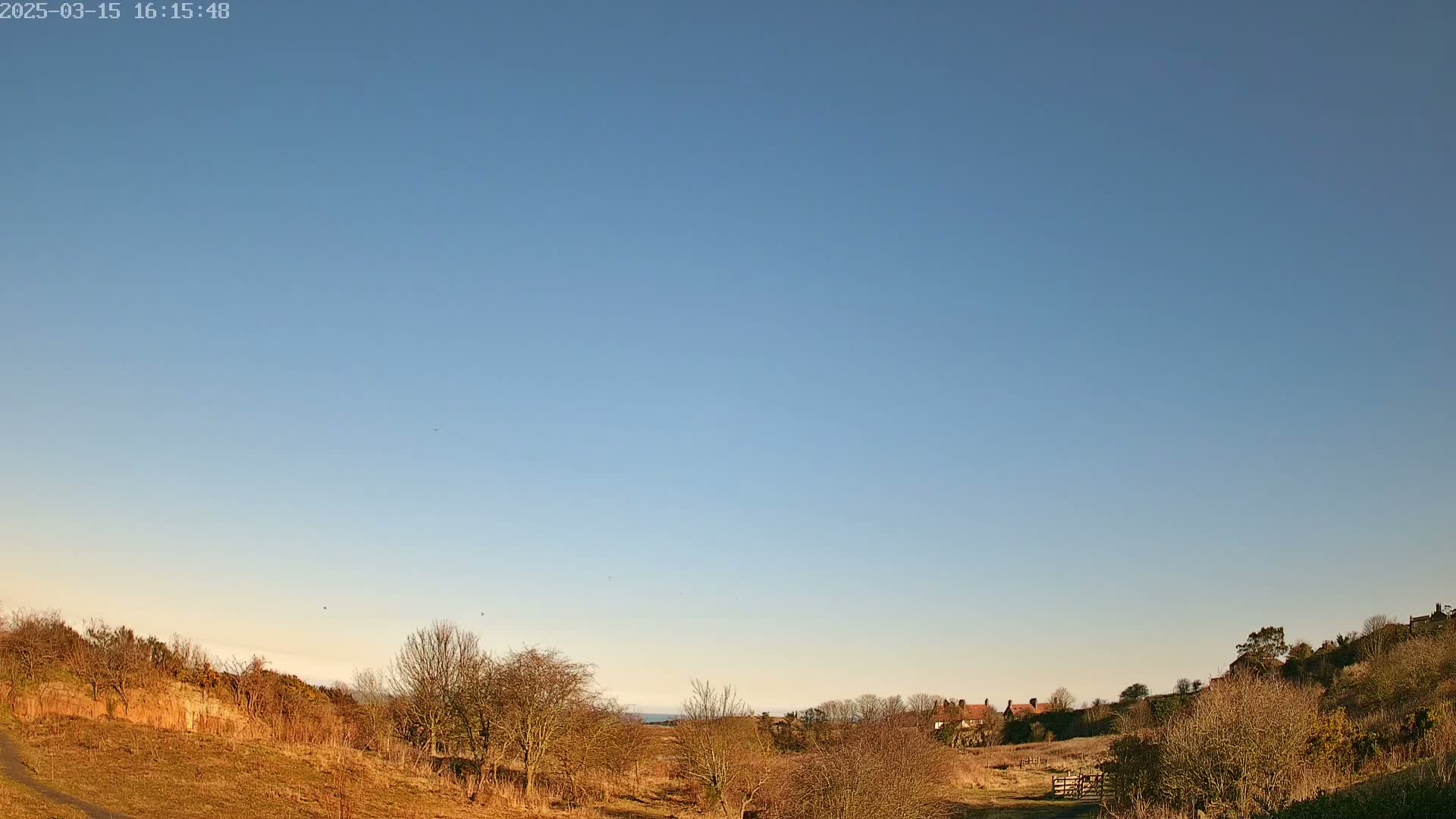 Under a clear blue sky, a hilly landscape of dormant, brown vegetation reveals a distant cluster of buildings.