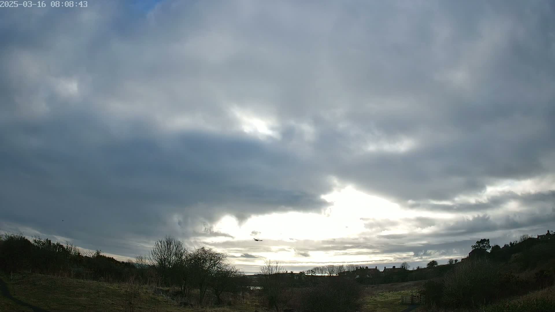 A mostly cloudy sky with patches of sunlight illuminates a landscape of low-lying shrubs and distant houses.