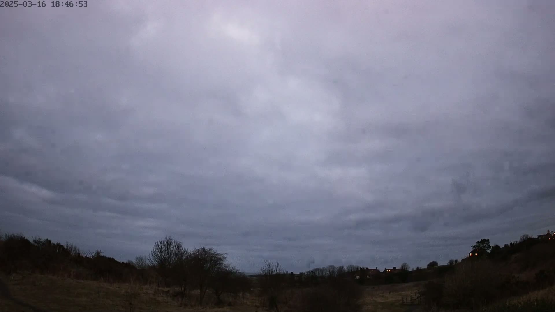 A mostly cloudy, dusky sky hangs over a sparsely vegetated landscape with a few houses visible in the distance.