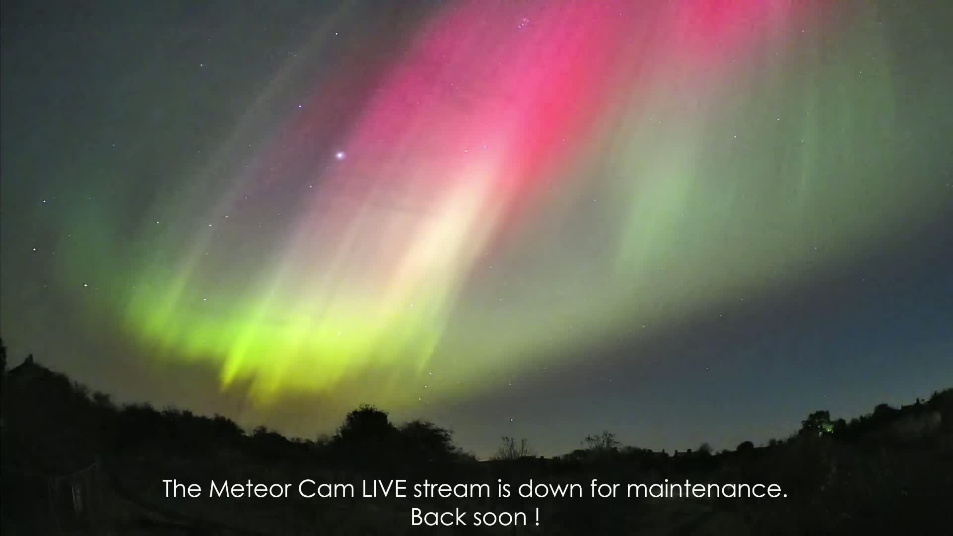 A vibrant aurora borealis, displaying shades of green and pink, illuminates the night sky above a dark, silhouetted landscape.