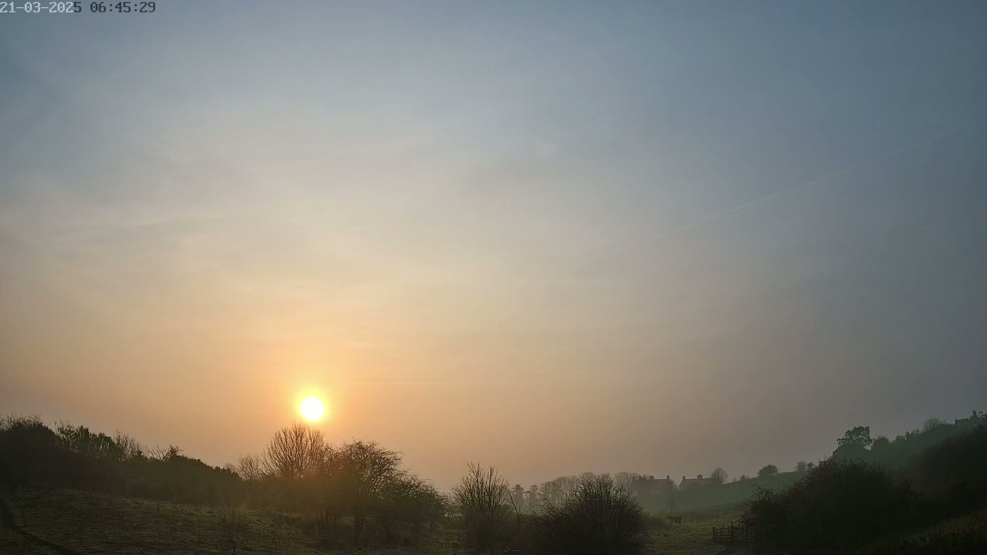 A hazy sunrise casts a warm glow over a rural landscape of low hills, trees, and distant buildings.