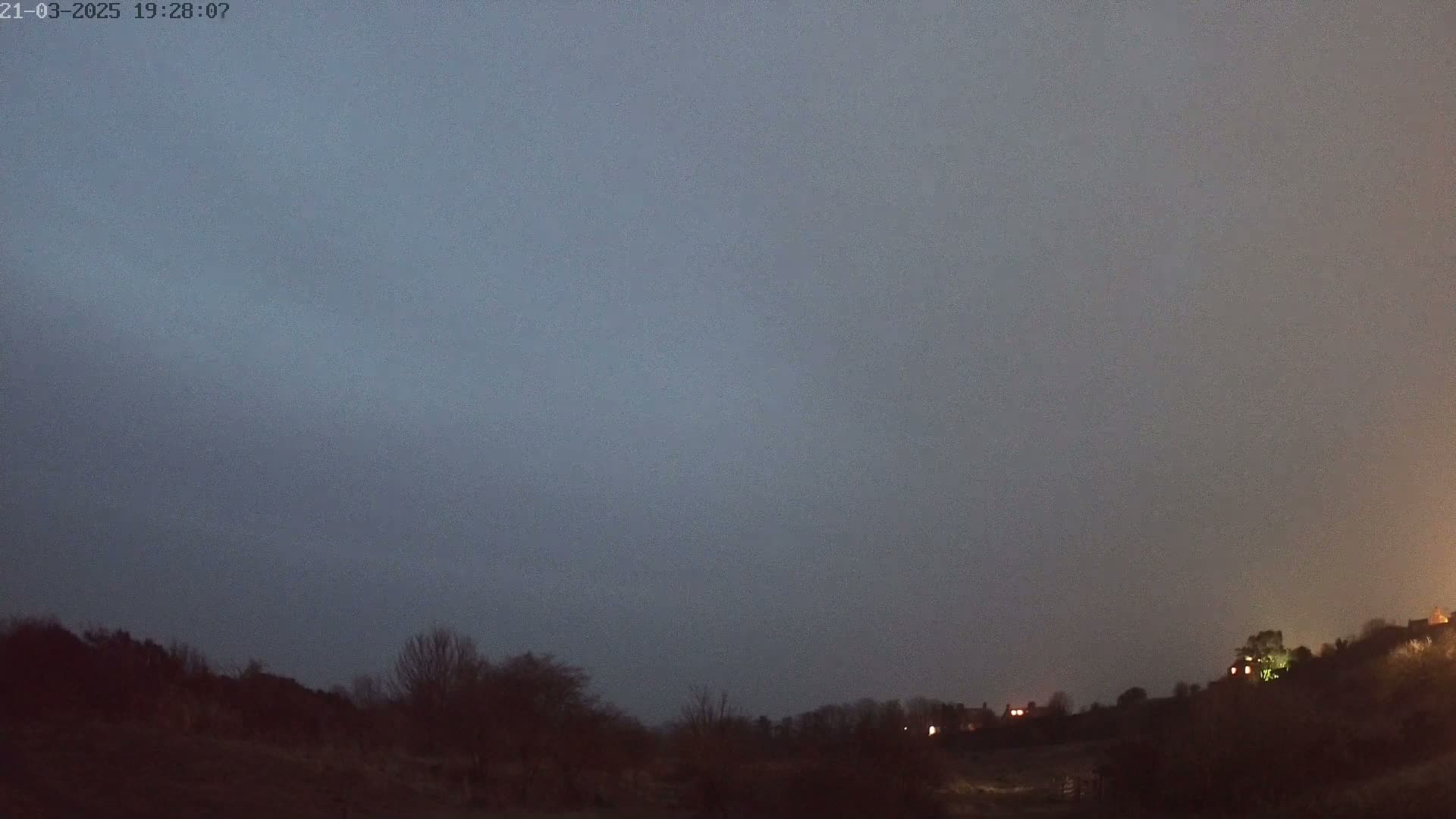 A dark, overcast sky hangs over a sparsely populated, hilly landscape at night, with a few faint lights visible in the distance.