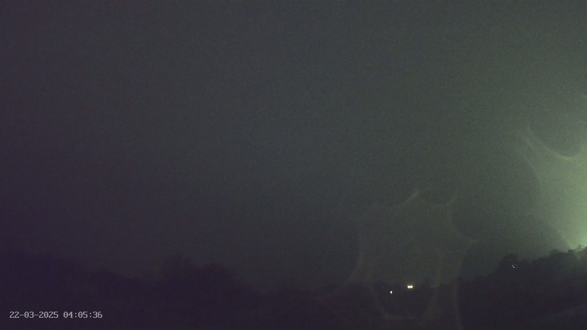 A dark, overcast night scene shows a distant line of trees and faint lights under a stormy sky.