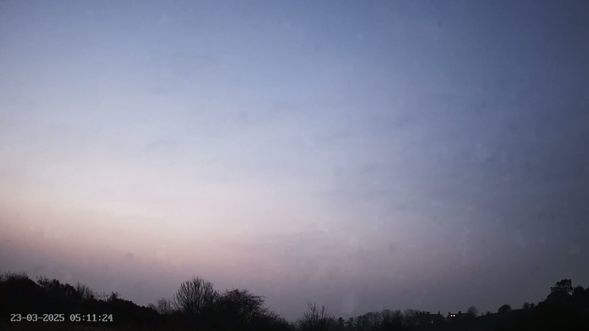A hazy, pale pink and purple dawn sky sits above a silhouetted landscape of trees and distant buildings.