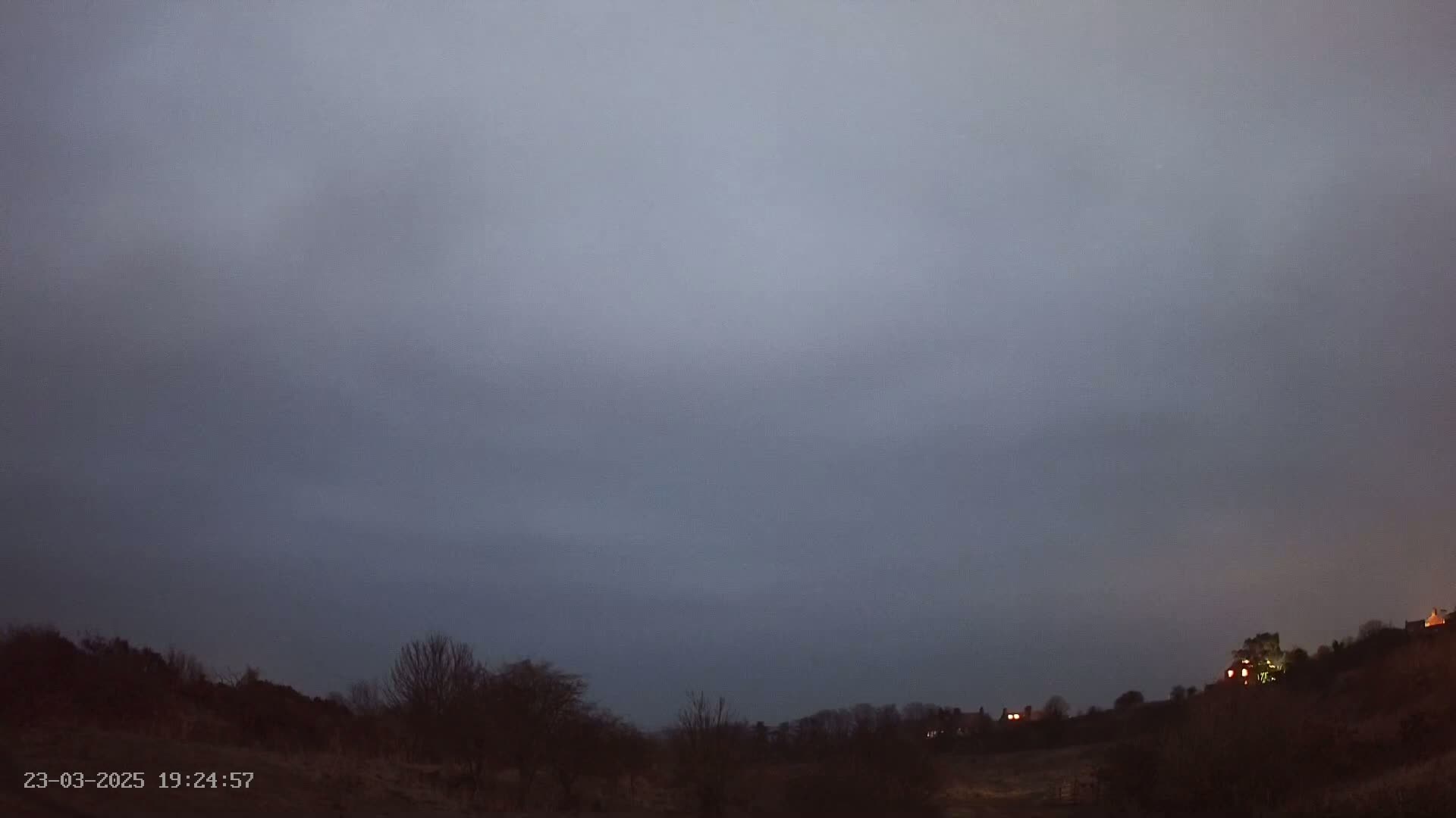 A dark, overcast sky hangs over a sparsely treed landscape at dusk, with a few distant lights visible on a hill.