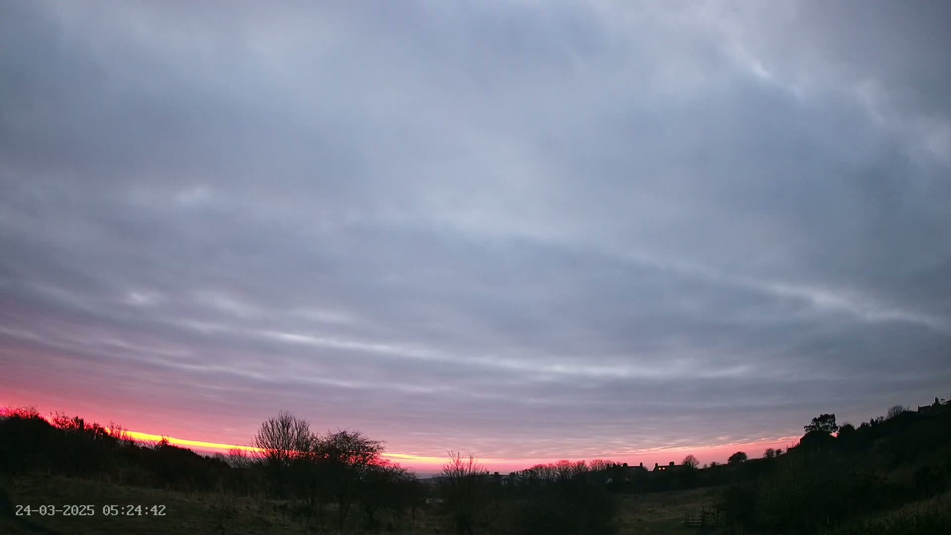 A pinkish-orange sunrise with thin clouds stretches across a mostly cloudy sky above a silhouetted landscape of trees and distant buildings.