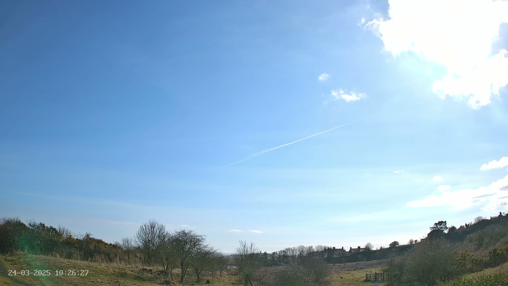 A mostly sunny sky with a few clouds and a contrail over a flat, grassy landscape with bare trees and houses in the distance.