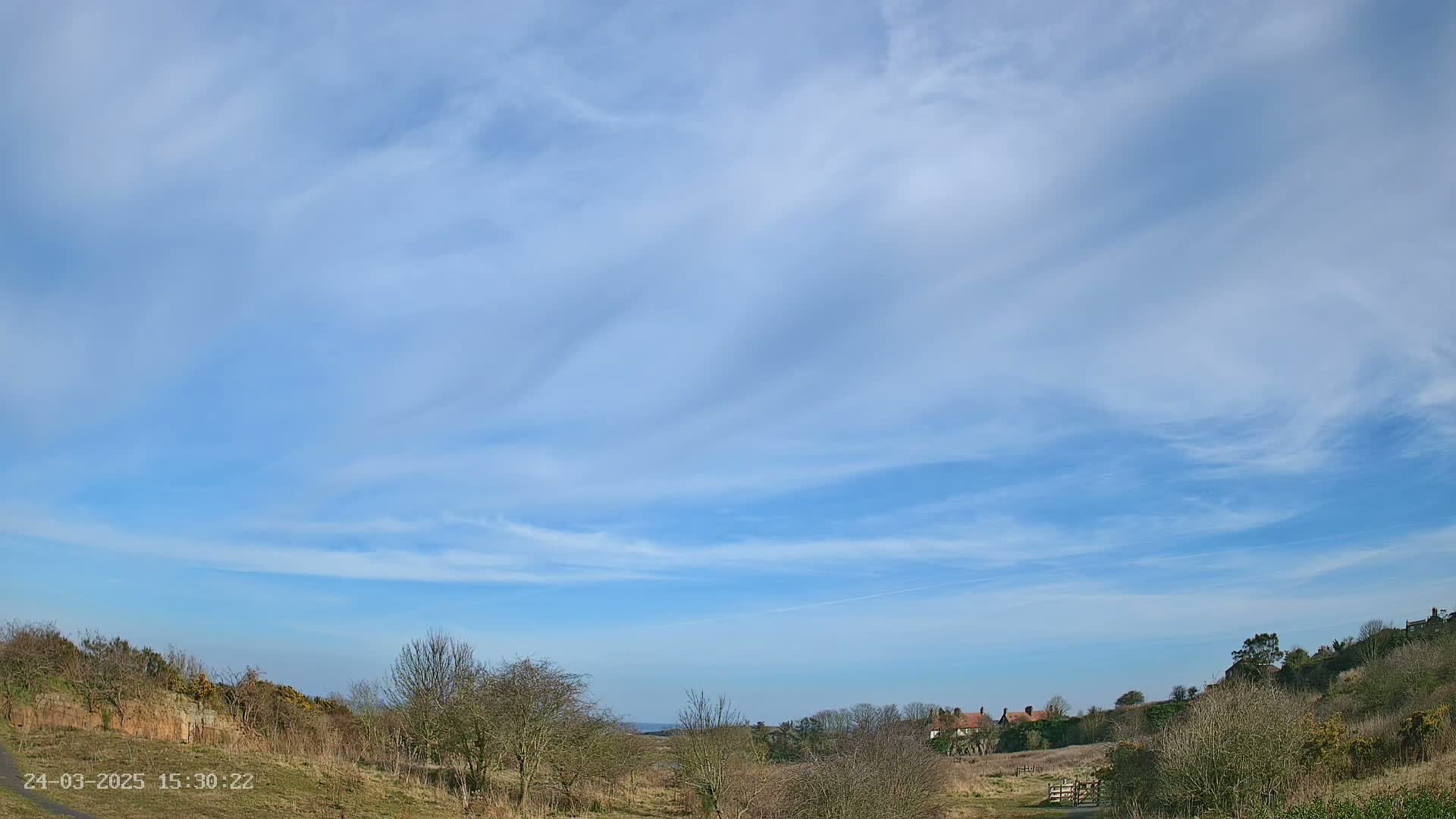 A mostly sunny day with wispy clouds overhead shows a view of a hilly landscape with sparse, leafless trees and a few distant houses.