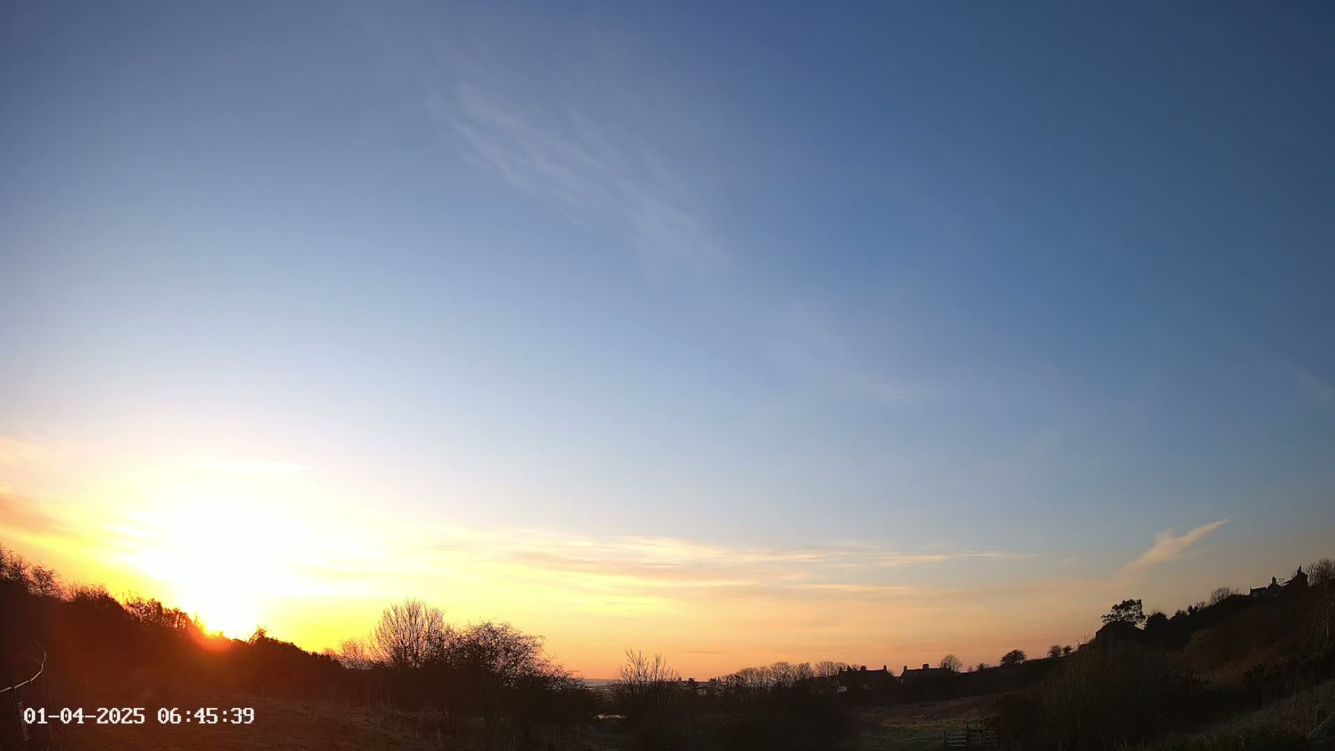 A vibrant sunrise casts a warm glow over a landscape of silhouetted trees and distant buildings under a mostly clear sky.