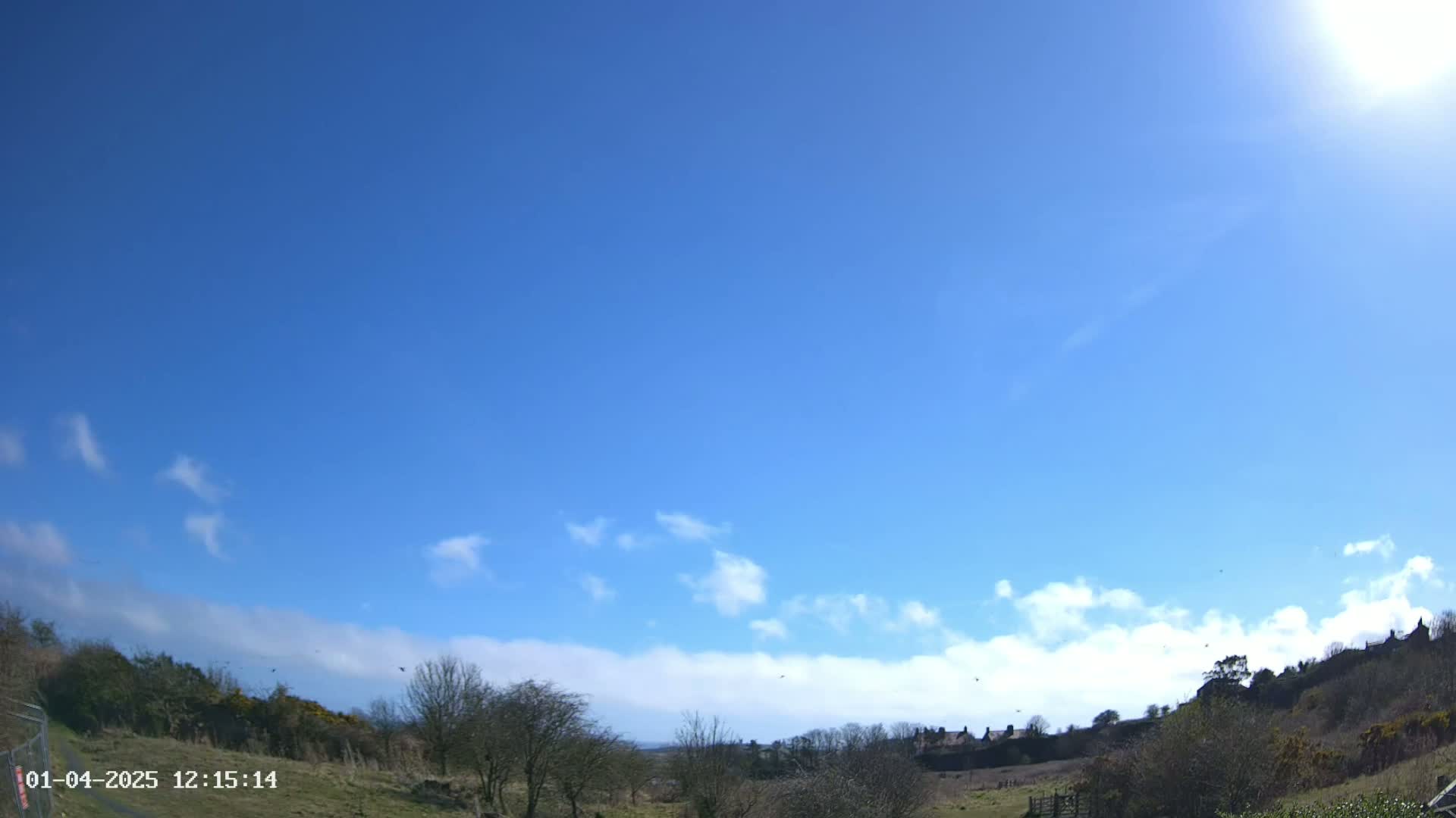 A sunny day with a mostly clear blue sky and a few scattered clouds, overlooking a landscape of low-lying hills, shrubs, and a few distant buildings.